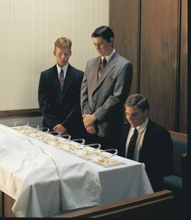 Two young men in suits stand near a sacrament table with their heads bowed while a third offers the prayer for the sacrament bread.