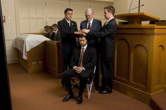 A young man in a black suit is given the gift of the Holy Ghost by three other men in a sacrament meeting setting.
