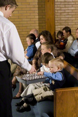 A small boy with blond hair reaches out to take a cup from a metal sacrament tray that a deacon is holding out to him.