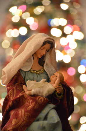 A Nativity figurine of Mary holding the baby Jesus wrapped in white linen, with colorful Christmas lights in the background.