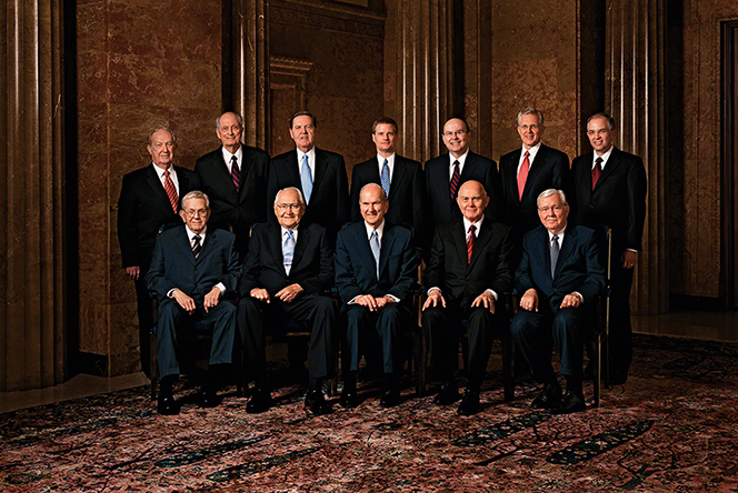 A portrait of the Quorum of the Twelve Apostles, five of them sitting in a row of chairs and the remaining seven standing behind them.