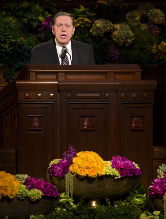 Elder Holland in a striped tie and black suit, standing and speaking behind the pulpit in general conference.