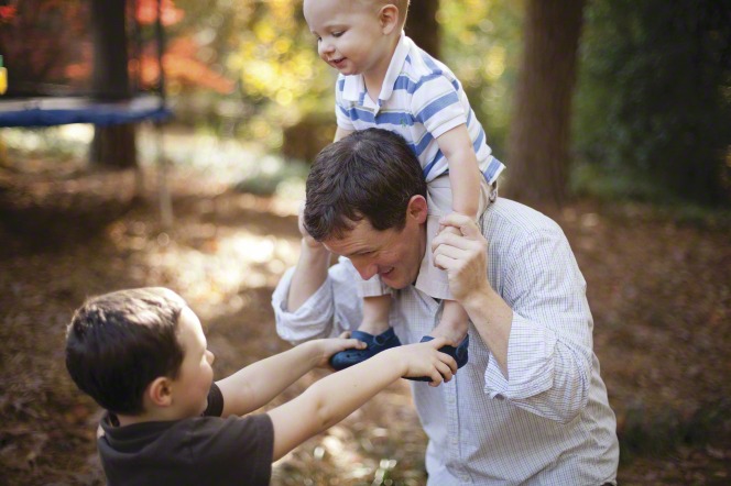 A father plays with two sons, one on his shoulders, in the park.