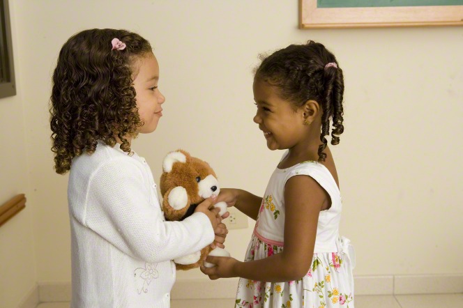 Two toddler girls in dresses standing and holding a teddy bear.