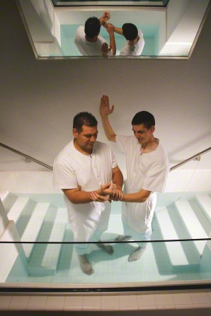 An elder missionary in white baptizes by raising his right arm and holding the wrist of another man in white in the baptismal font.