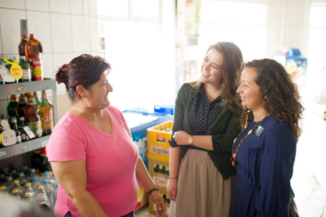 Two sister missionaries from Romania standing in a store, smiling and talking to a woman in a pink shirt.