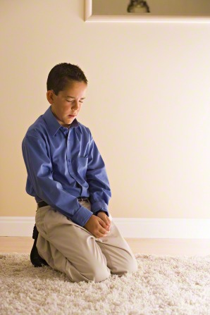 A boy kneels on a rug, clasps his hands, and prays.