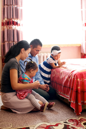A family kneel in a bedroom next to a bed and pray together.
