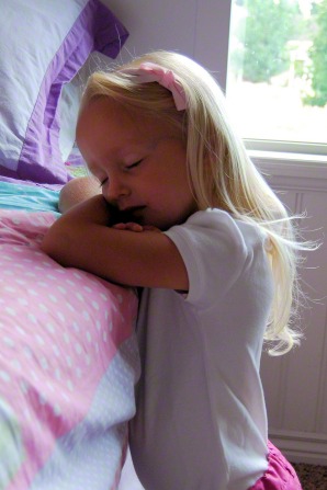 A girl wearing a white T-shirt and a pink bow in her blond hair kneels next to her bed with her eyes closed to say a prayer in the morning.
