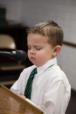 A little boy closes his eyes and folds his arms while he prays at a microphone in Primary.