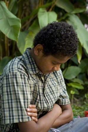 A young man with curly black hair and a green plaid shirt sits in an outdoor tropical setting and prays with head bowed and arms folded.