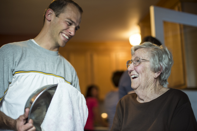 A young man smiles while drying a dish in a kitchen and looking toward an elderly woman whom he is serving.