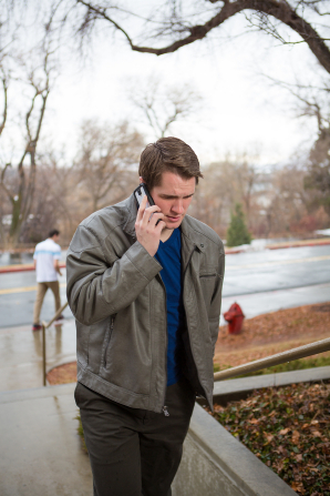 A young man in a gray jacket walks outside while holding a phone close to his ear.