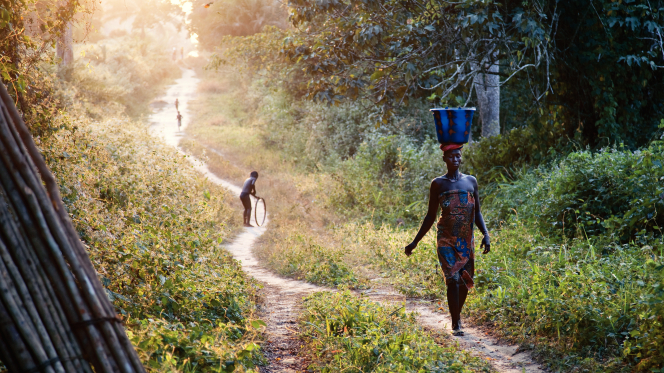 An African woman balancing a large bucket of water on her head while walking down a narrow path.