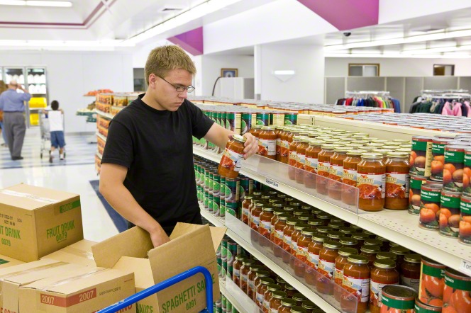 A man in a black shirt lifting food jars out of cardboard boxes onto shelves at the bishops’ storehouse.