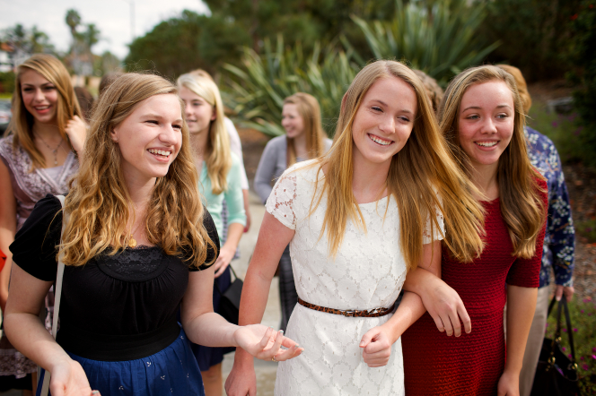 A group of young women link arms and walk together outside, smiling.