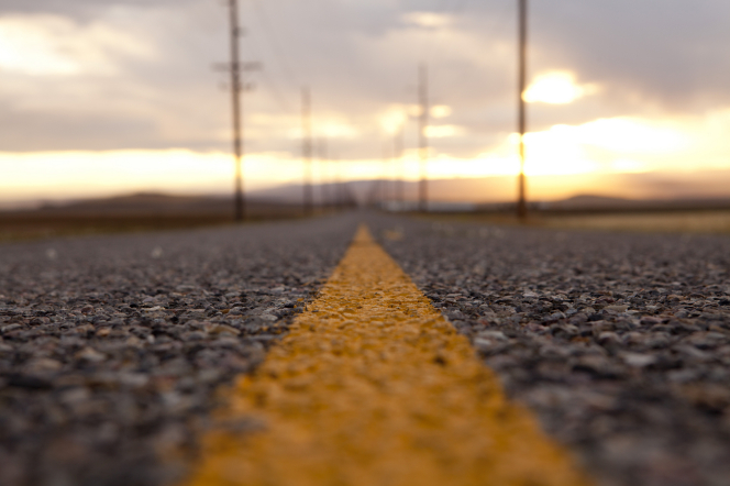 A detailed view of the yellow painted line running down the middle of an asphalt road, blurring out in the background toward a sunset on the horizon.