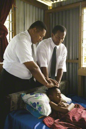 Two men in white shirts and ties give a priesthood blessing to a young boy who is lying sick in bed.