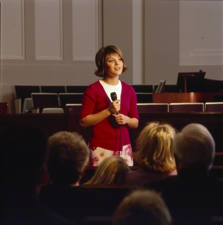 A woman is standing in the middle of a congregation, holding a microphone and bearing her testimony.