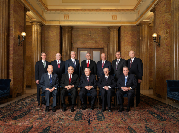 A portrait of the Quorum of the Twelve Apostles, five of them sitting in a row of chairs and the remaining seven standing behind them.