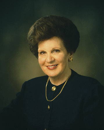 A photograph of Mary Ellen Wood Smoot against a gray background, wearing a gold necklace and a black dress with gold buttons down the front.
