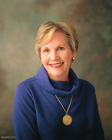 A photograph of Bonnie Dansie Parkin against a gray and brown background, wearing a blue blouse and a gold necklace with a pendant.