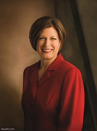 A photograph of Julie Bangerter Beck against a brown background, wearing a red blazer.