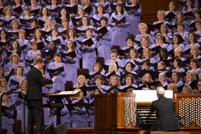 Ryan Murphy in a black suit stands in front of the women of the Mormon Tabernacle Choir to conduct a song.