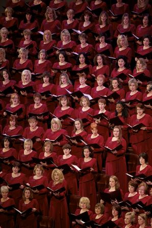 Rows of women in bright red dresses hold their music in folders and sing in the Conference Center with the rest of the Mormon Tabernacle Choir.