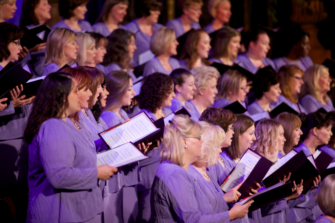 A section of women from the Mormon Tabernacle Choir seen from the side, holding their music in folders and singing.
