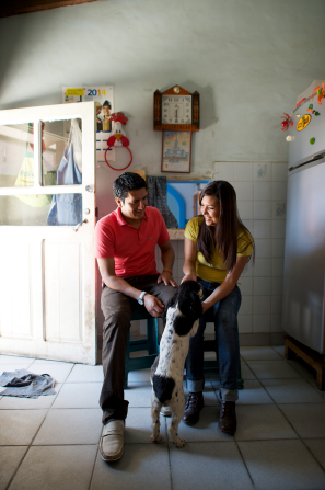 A young couple from Bolivia sitting on wooden stools while a dog stands on two feet and rests the other two feet on the woman’s leg.