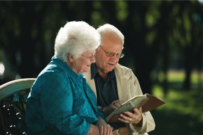 An elderly couple sits on a bench in the park and reads the scriptures together.