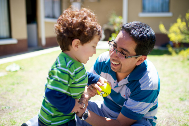 A man plays outside with his curly-haired toddler, who is reaching for a small yellow ball and laughing.