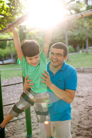 A man in a blue shirt holds his son up on some handlebars on a playground at a park.
