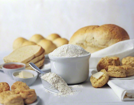 A setting of flour in a bowl and butter and jam in a bowl, with sliced bread, a loaf of bread, rolls, and biscuits on plates.