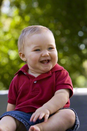 A portrait of a baby boy in a red shirt, sitting outside and smiling.