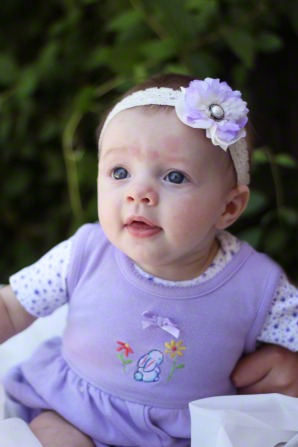 A baby girl in a purple dress and a flower headband, sitting and smiling.
