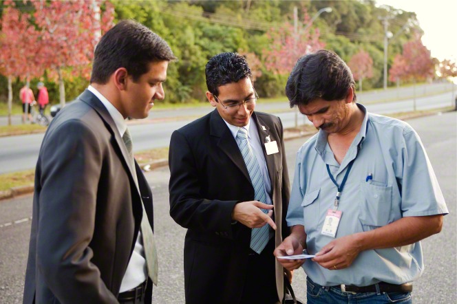 Two elder missionaries in suits standing and giving a pass-along card to a man in a blue shirt in Brazil.