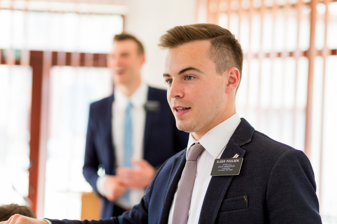 An elder missionary with brown hair, a white shirt, a gray tie, and a blue suit, standing in a classroom with his companion in the background.