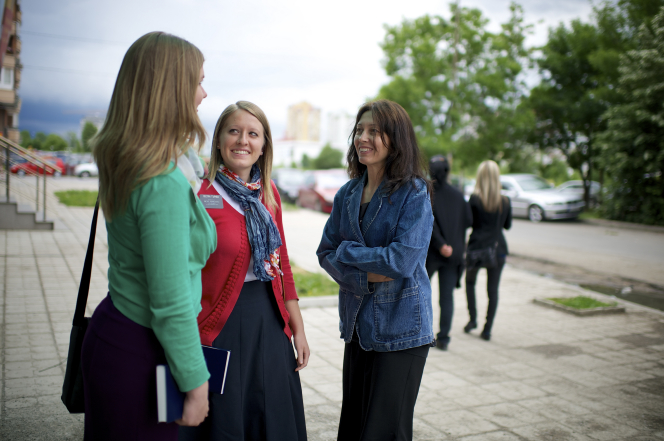 A woman with brown hair, a black dress, and denim jacket talks to two sister missionaries outside in Bulgaria.