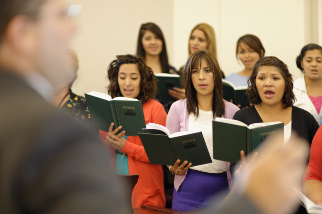 A ward choir consisting of several women, holding hymnbooks and singing in a chapel.