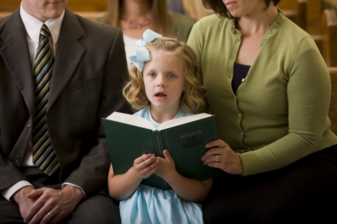 A young girl with a bow in her hair and a light blue dress sits between her parents while singing from a hymnbook.
