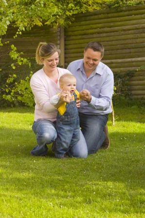 A mother and father kneel in the grass next to their son and help him while he tries to walk.