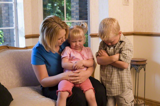 A mother sits on a couch with her daughter in her lap and her son next to her while all three pray.