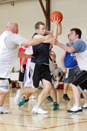 A man playing basketball with a group of men in a gym, holding the ball high.