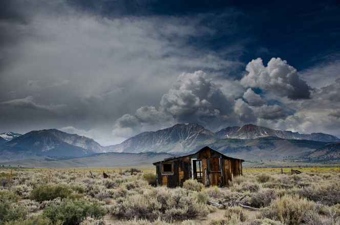 A run-down cabin in a field, with a mountain range covered with clouds in the background.