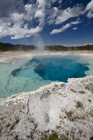 Hot springs by a hillside at Yellowstone National Park, with clouds in the clear, blue sky.