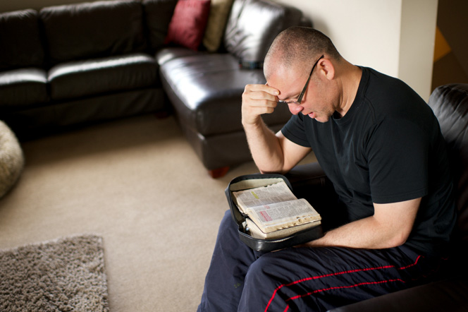 A man sits in an armchair with his scriptures in his lap and reads from them.