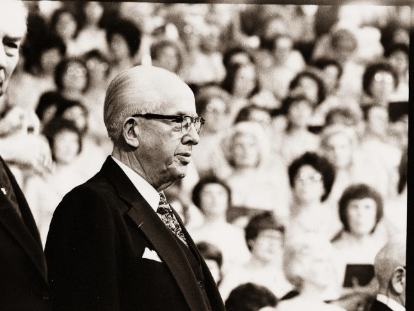 President Benson in a suit, a white shirt, and glasses, standing and speaking to the audience at general conference.