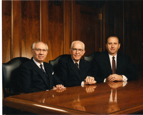 President Ezra Taft Benson sitting at a table between his two counselors in the First Presidency, President Gordon B. Hinckley and President Thomas S. Monson.